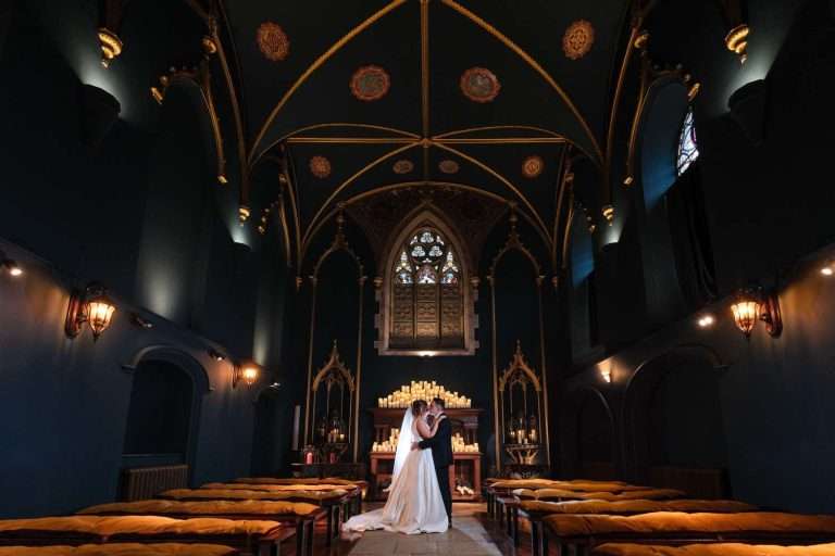 A couple in formal attire stands at the altar in a beautifully decorated, dimly lit, Gothic-style chapel with a high vaulted ceiling and numerous candles—perfect for capturing timeless Lartington Hall wedding photos.