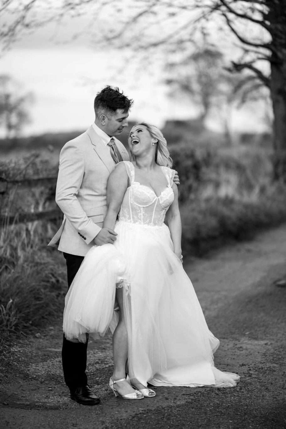 A couple laughs together on a rural path. The woman in a wedding dress lifts her skirt slightly, while the man in a light suit stands beside her. Trees and a fence line the background.