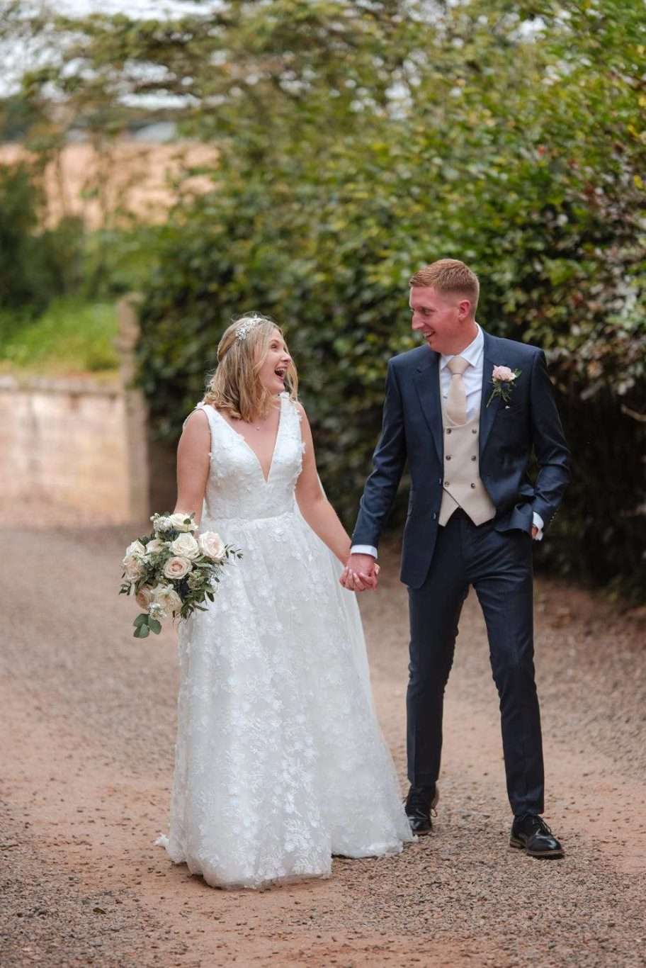 A bride and groom hold hands and smile at each other while walking on a gravel path. The bride is wearing a white dress and holding a bouquet, and the groom is in a suit.