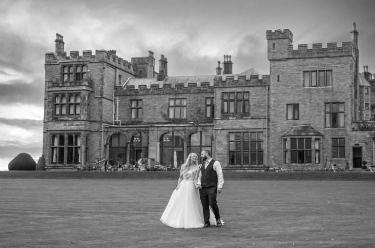 A bride and groom stand together on a lawn in front of a large historic stone building, posing for a wedding photo—perfect for documentary Lake District wedding photography or fun and relaxed highlight films.