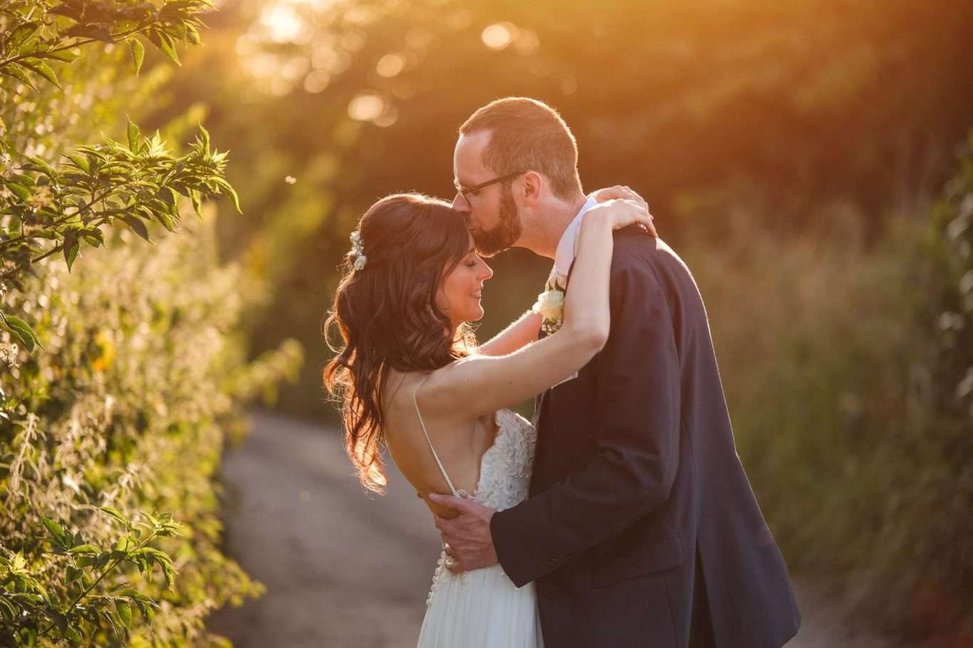 A bride and groom embrace outdoors on a sunlit path, with the groom kissing the bride’s forehead and greenery in the background, beautifully captured by a North East Wedding Photographer.