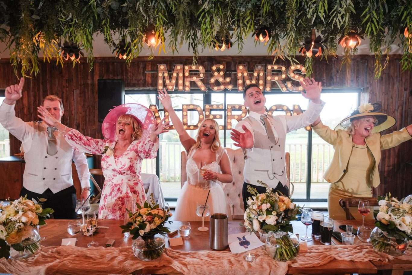 Five people in formal attire sit and stand behind a decorated table at a wedding reception, cheering with raised arms; a lit "MR & MRS" sign glows behind them, perfectly captured by a North East Wedding Photographer.