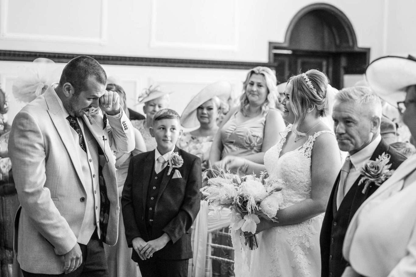 A groom wipes his eyes as he stands beside the bride during a wedding ceremony, surrounded by guests and family in formal attire, beautifully captured by a North East Wedding Photographer.