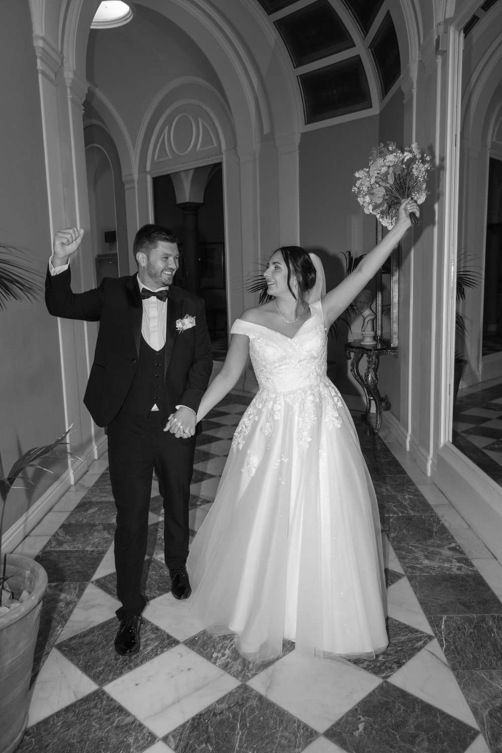 A bride in a wedding dress and groom in a suit hold hands and celebrate whilst walking in an elegant corridor—captured by a Newcastle wedding photographer using hybrid wedding photography for timeless memories.