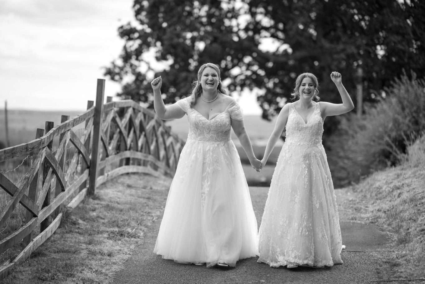 Two women in wedding dresses walk hand in hand on a path outdoors, smiling and raising their free arms in celebration.