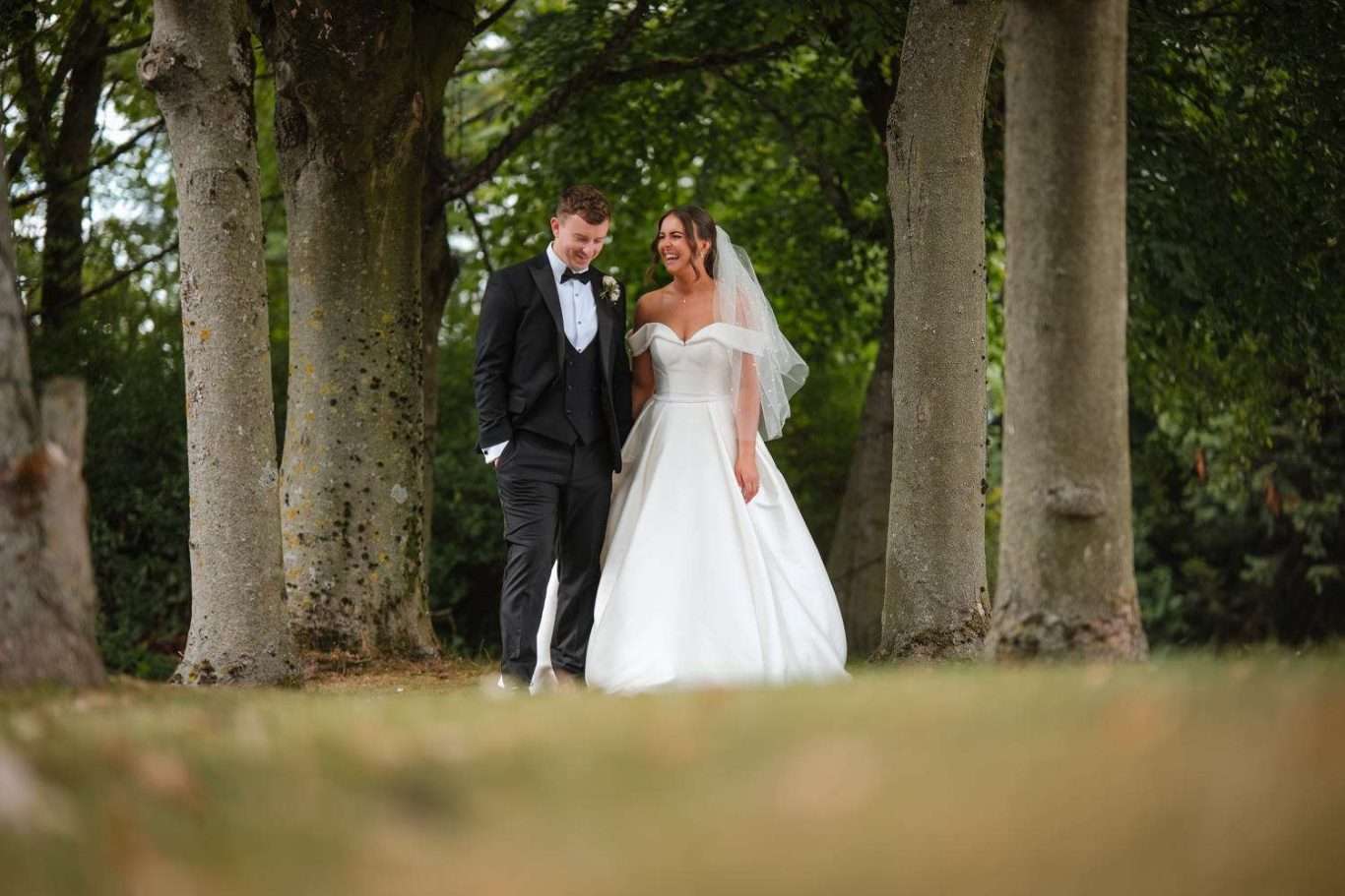 A bride and groom in formal wedding attire walk together outdoors, surrounded by tall trees and greenery, beautifully captured in timeless, natural wedding photos by a Newcastle photographer.